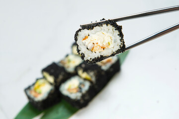 Close up of Sushi Roll piece wrapped in rice and Black flying fish roe (Tobiko caviar) hold in metal chopsticks with blurred Japanese dish with avocado, unagi eel inside on white marble background
