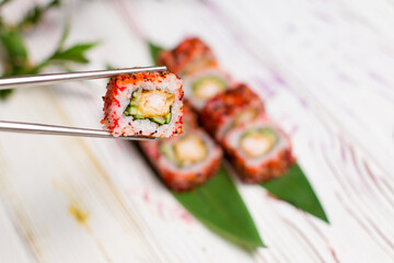 Close up of Sushi Roll piece wrapped in rice and flying fish roe in metal chopsticks with blurred background. Sushi Roll with tempura shrimp inside on bamboo leaves and plant on background. Copy space