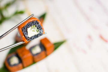 Close up of Sushi Roll piece with black rice and salmon on top in metal chopsticks with blurred background. Philadelphia Sushi Roll with bamboo leaves and plant on background. Copy space.
