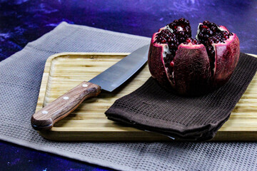 An open pomegranate fruit that is divided into five parts that are held together on a napkin / cloth on a wooden tray on a blue background. Large red ripe pomegranate with a knife next to it.