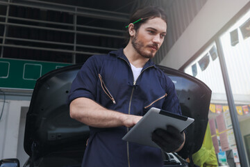 Auto mechanic holding digital tablet and working at garage, car service technician checking and repairing customer’s car at automobile service center, vehicle repair service shop concept