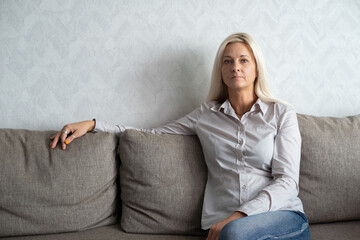 A confident female businesswoman sits on a gray sofa against the background of wallpaper and looks...