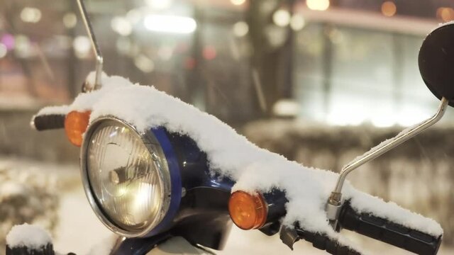 The steering wheel and headlight of a moped covered with snow during a cyclone on a city street in the evening. Close-up. Urban winter landscape
