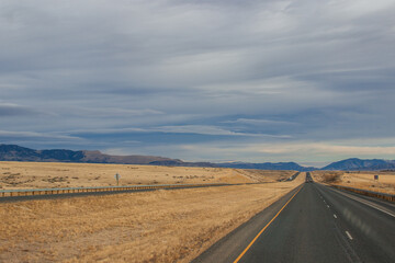 Asphalt road among steppes with yellow dry grass, blue mountains on the horizon, beautiful blue-gray autumn sky. Montana, USA, 11-23-2019