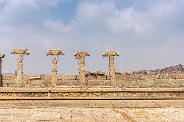 Fototapeta premium Hampi, Karnataka, India - November 5, 2013: Vijaya Vitthala Temple. Remains of brown stone wall pillars with missing roof under blue cloudscape,