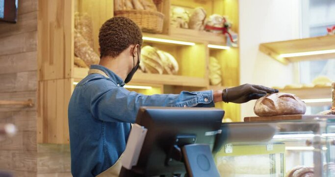 Young African-American bakery saller starts work day and spreads fresh bread on counter in protective mask and gloves.