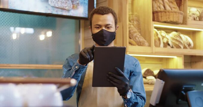 Young African American bakery worker stands on background of shelves with bread in protective mask and makes ordering products online using tablet.