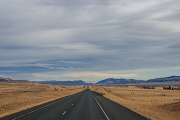 Asphalt road among steppes with yellow dry grass, blue mountains on the horizon, beautiful blue-gray autumn sky. Montana, USA, 11-23-2019