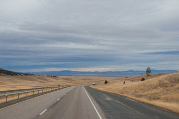 Fototapeta na wymiar Asphalt road among steppes with yellow dry grass, blue mountains on the horizon, beautiful blue-gray autumn sky. Montana, USA, 11-23-2019
