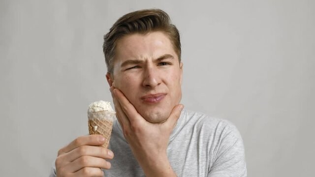 Young guy eating ice-cream cone and suffering from acute toothpain, grey studio background, close up portrait