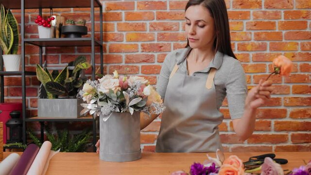Portrait young woman florist in apron at workplace in flower shop. Brunette girl making bunch of flowers in flowers box. Smiling female florist professional makes bouquet. Composition in hatbox.