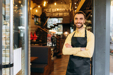 Portrait of an attractive bearded caucasian male waiter, barista or owner of a restaurant, cafe or bar stands at the entrance wearing a protective mask and a black work apron with crossed arms and