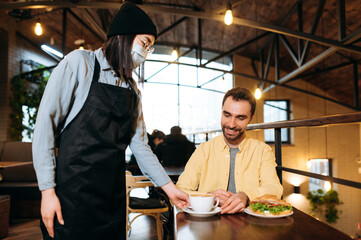 A waiter girl in a protective medical mask and a black apron brought aromatic coffee to a guest of restaurant or cafe shop. Young caucasian guy with a smile got his coffee while sitting in a cafe