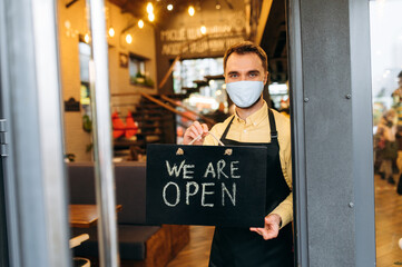 We are OPEN. Portrait of a satisfied caucasian waiter or barista standing at restaurant or cafe entrance wearing medical mask on his face and black apron. Employee man showing signboard OPEN in his