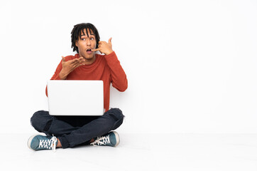 Young african american man sitting on the floor and working with his laptop making phone gesture and doubting