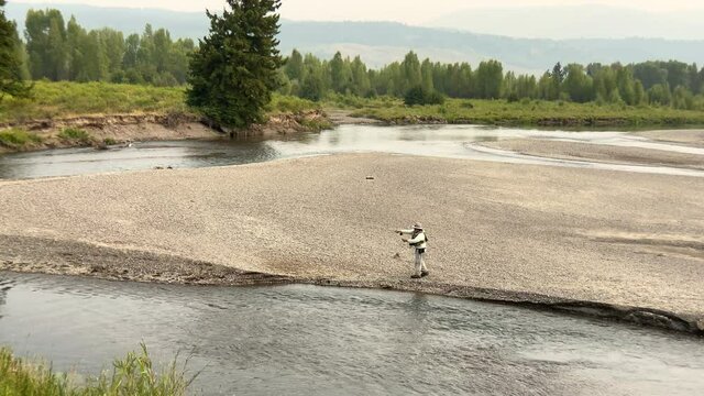 Fisherman fly fishing from a sandbank in the Buffalo Fork River with forested hills in the distance, Bridger-Teton National Forest, Wyoming. Wide static shot with slight panorama.