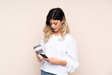 Fototapeta premium Teenager girl isolated on beige background holding coffee to take away and a mobile