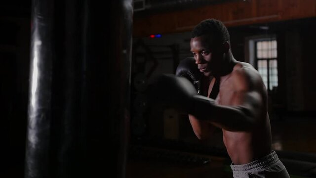 African-american athletic half-naked young man boxer training punching on the punching bag in the gym