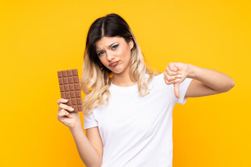 Teenager girl isolated on  yellow background taking a chocolate tablet making bad signal