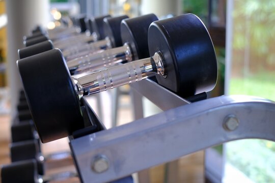 A close-up picture of steel dumbbells with black rubber protection lining up in rows on a metal rack in a gym by a glass window.