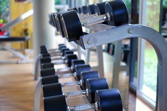 A close-up picture of steel dumbbells with black rubber protection lining up in rows on a metal rack in a gym by a glass window.