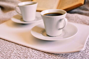 Pair of coffee cups with hot black coffee on white tray on cloth and open book on blurred background
