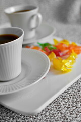 Pair of coffee cups with hot black coffee and candies in colorful wrappers on white tray on cloth close up. Tilt horizon. Selective focus on first cup