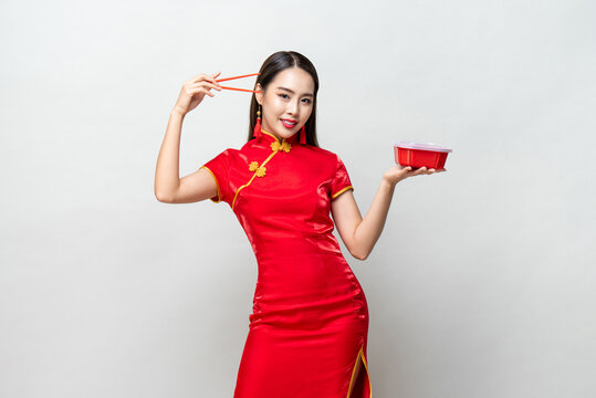 Pretty Asian woman in traditional red Chinese qipao dress holding plastic food box and chopsticks in studio isolated gray background