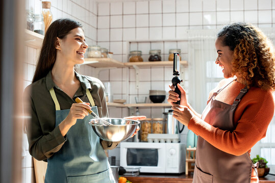 Happy two woman friends shooting video with mobile phone to share online while cooking in kitchen at home