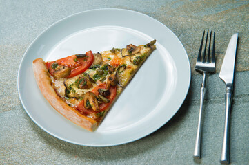 Slice of veggie pizza on gray plate. Knife and fork near the dish on stone anthracite color background. Piece of vegetable pizza with tomatoes, champignon mushrooms, sliced eggplant with pesto sauce
