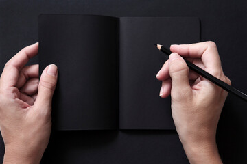 Manos escribiendo en una libreta negra con un lápiz negro sobre un fondo negro. Espacio libre para texto. - obrazy, fototapety, plakaty
