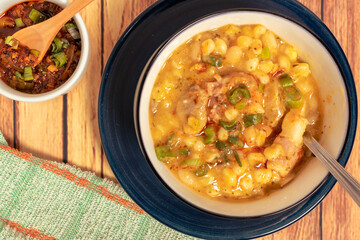 White earthenware bowl on a blue rimmed plate with Locro typical of Argentine cuisine and the spicy sauce that accompanies this stew on a wooden table. aerial view.