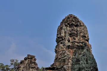 The top of the famous Bayon Temple, Angkor against the blue sky. Huge human faces, carved from stone, look in all directions. The tower is weathered, damage from time is visible. UNESCO. Cambodia