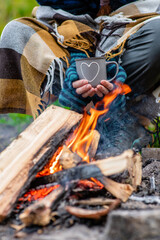 tourist wrapped a plaid holds cup of the hot tea  near a bonfire