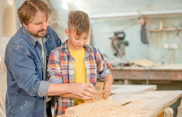 Carpenter teaches young boy to plan wood in a carpentry workshop. Empty space for text