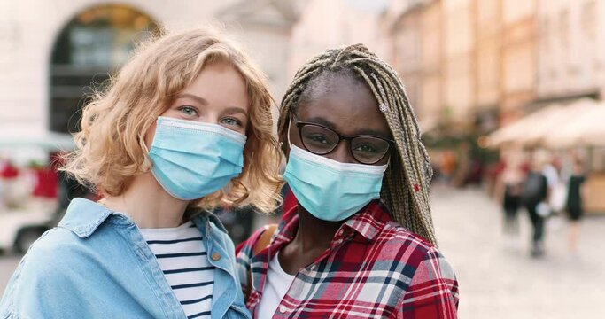 Portrait of young happy mixed-races females best friends in medical masks standing outdoor in city street and looking at camera. Multi ethnic women in respiratory protection posing to camera in town.
