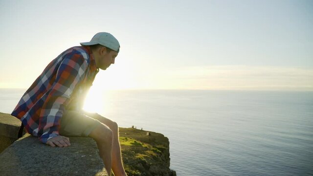 Young man in shirt, shorts and cap sitting on the edge of a cliff looking at sunset view, ocean and sunlight in the background, Madeira, Portugal