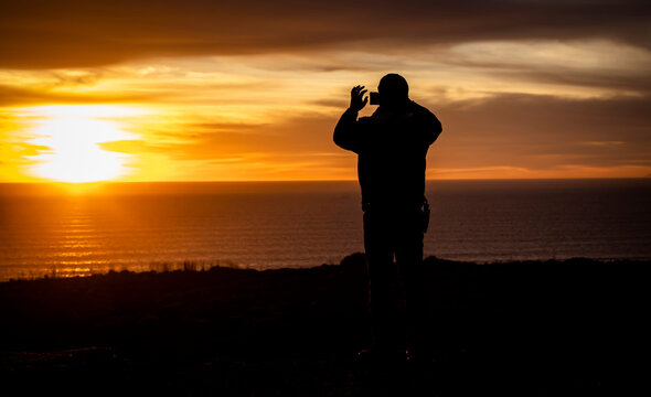 Silhouette of a man looking out at a sunset or sunrise taking a cell phone picture