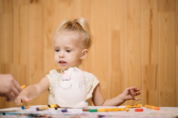 Little girl paints a piggy bank with markers