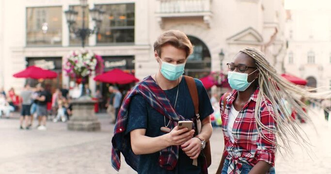Mixed-races couple or male and female friends in medical masks walking in city street and looking at mobile phone. Young multi ethnic man and woman using smartphone and talking. Quarantine.