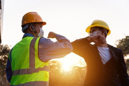 Asian industry construction site worker and foreman wearing hygiene face mask elbow bump greeting adaptation to prevent Coronavirus or Covid-19 spreading at warehouse near the sunset, New Normal