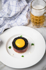 Gourmet Rindertatar Beef Tatar roh aus Filet fettarm, Kapern, Eigelb, Zwiebeln auf Toast Brot und schwarzer Kaviar mit Bier, Küchentuch und Marmor Hintergrund hell
