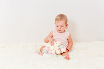 Cute European baby girl one year old sits on a white fluffy plaid and plays and marshmallows on a white background