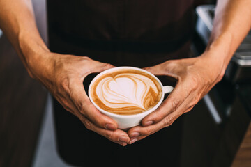 Barista hands holding a cup of hot heart-shaped latte art. Latte coffee with swirl milk decoration.