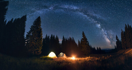 Panoramic view of night camping in valley with large pine trees. Burning campfire and illuminated...