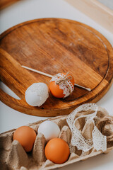 Easter eggs in carton box on white background. Happy Easter card. Easter eggs with lace border, wooden cutting board, brush on white table. Process of decorating easter eggs with lace and rope.
