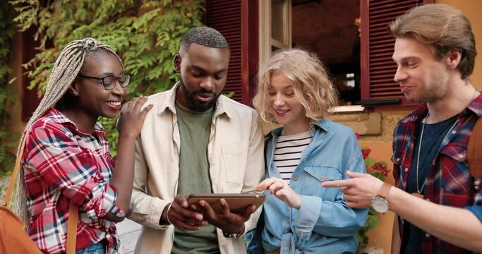 Mixed-races young males and females happy friends standing outdoors and looking at tablet device screen while tapping and scrolling. Multi ethnic people using gadget computer at street.