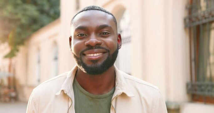 Portrait of handsome African American young man smiling joyfully to camera on sunny day in summer. Close up of happy face of cheerful guy outdoors in sunlight.
