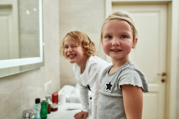 Portrait of cute little girl smiling at camera while washing her face, brushing teeth together with her sibling brother in the bathroom