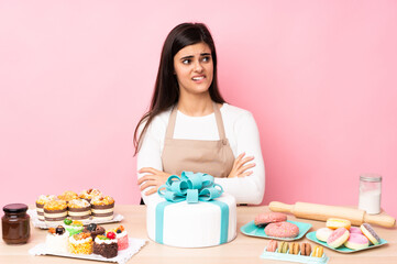Obraz na płótnie Canvas Pastry chef with a big cake in a table over isolated pink background with confuse face expression